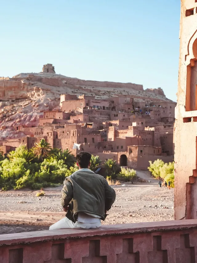 A traveller sitting on a kasbah wall looking across the palms to the Aït Ben Haddou ksar