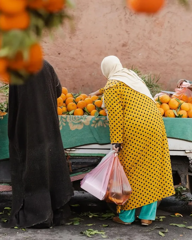 Two women choosing oranges from a market truck against a pink Marrakech wall